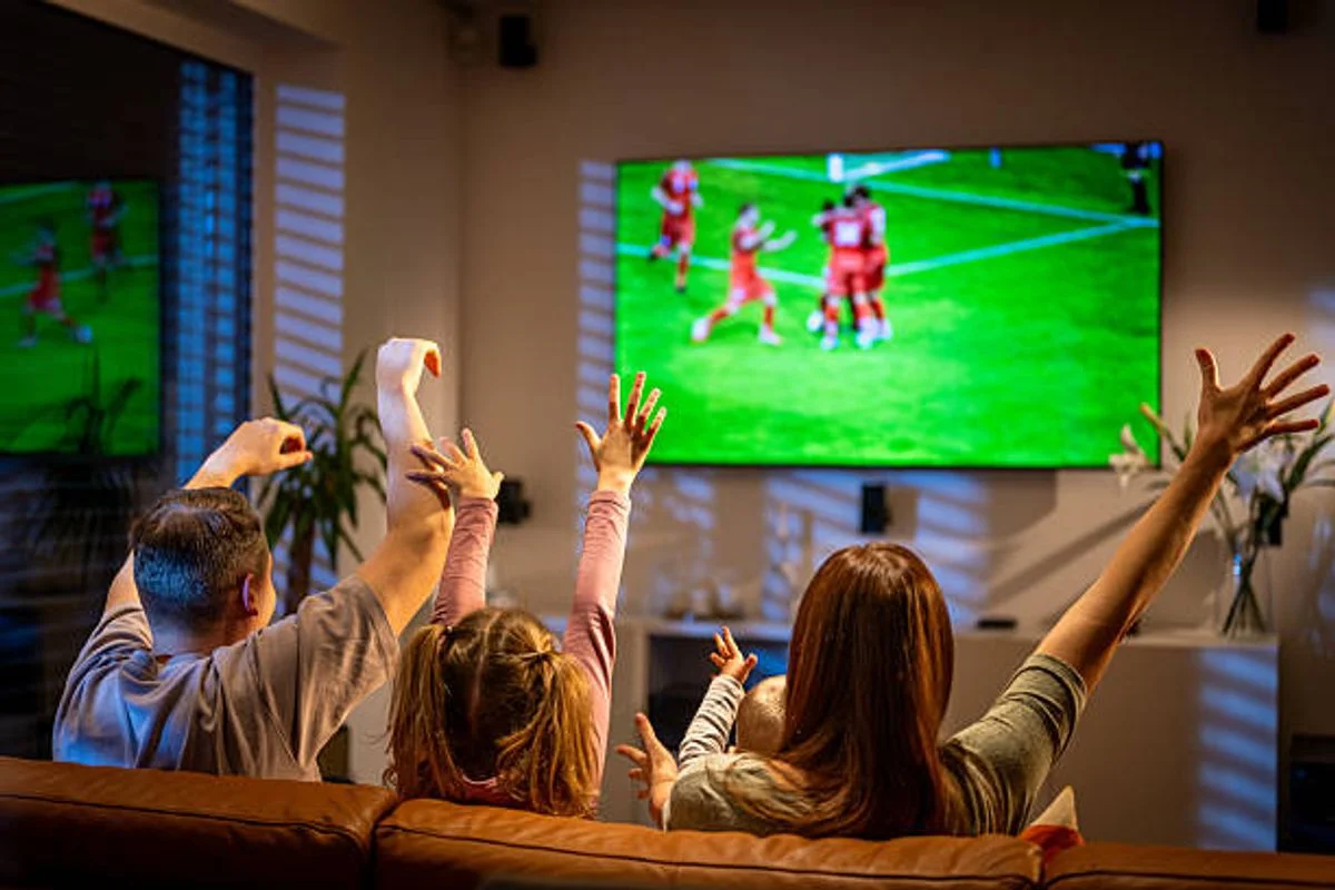 Candid shot of mid-aged parents and young daughter sitting on brown sofa, arms raised in celebration while watching soccer match on large-screen television.